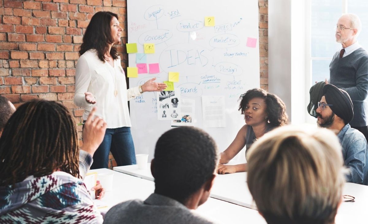 A group of people are meeting, one woman is standing and speaking to the group, one woman is making (side) eye contact with the camera